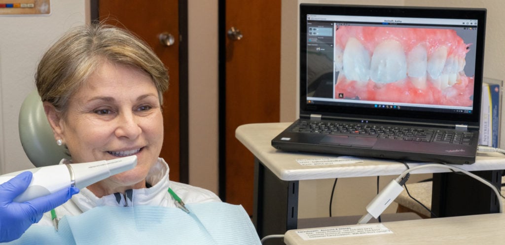 Woman undergoing a dental scan with an intraoral camera, with images displayed on a nearby laptop.