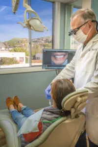Dentist examining a patient in a treatment room with a view of a hillside and dental images on a monitor.